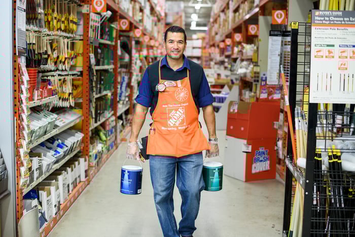 A worker wearing a Home Depot apron while holding two paint buckets in the aisle of a Home Depot store.