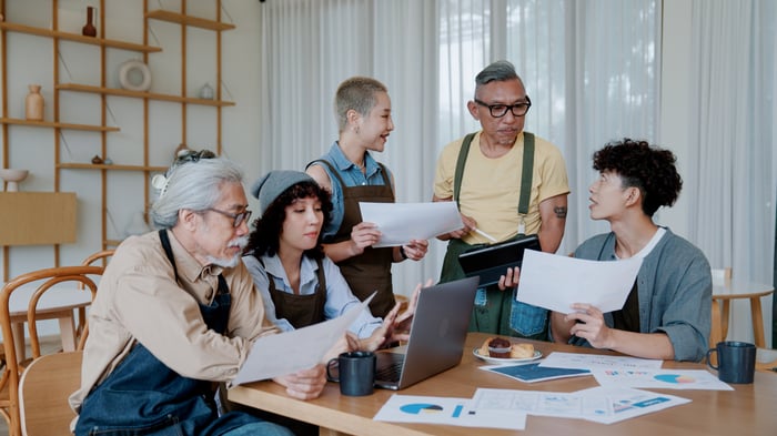 A group of investors sit and stand around a kitchen table referring to laptops and printouts.