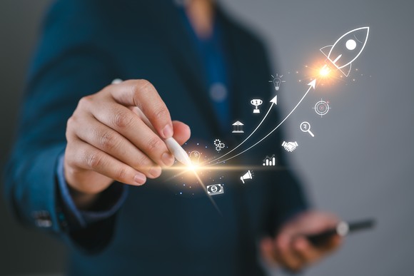 A person holding a pen to a digital image of a rocket, upward sloping lines, and small icons.