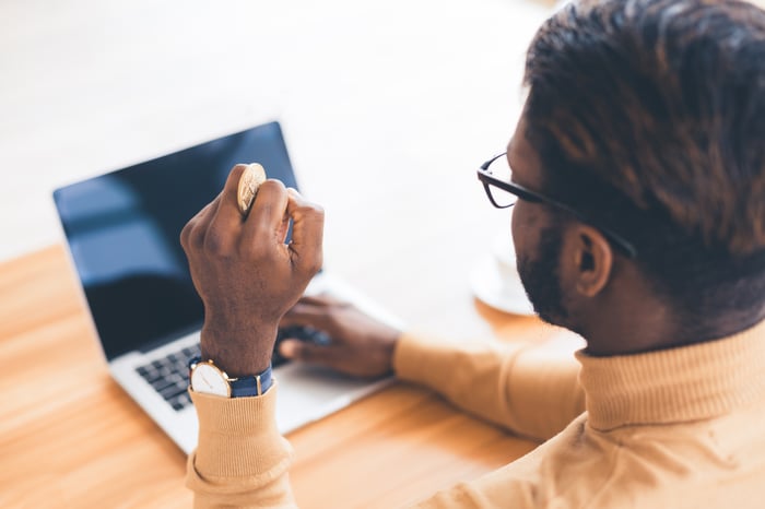 A person at their laptop holding a gold Bitcoin in their hand.
