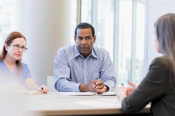 Three investors sit around a conference room table with some papers in front of them as two investors listen to the third speaking.