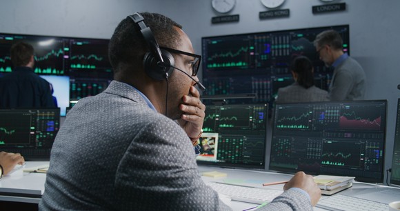 Person at desk in front of trading screens, with hand on chin.