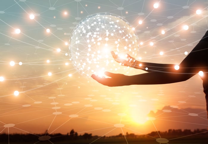 Silhouette of hands holding a sphere of interconnected dots of light against a sunset or sunrise crossed by orbs of light. 
