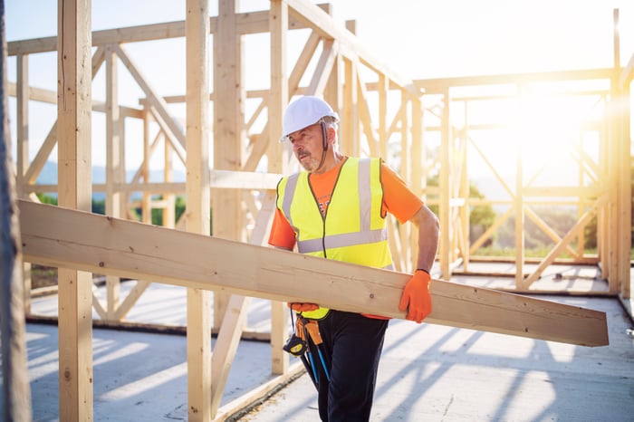 A person wearing a hard hat carrying a board at a house under construction.