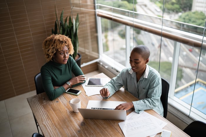 Two investors sit at a conference room table with a laptop, a tablet, a smart phone, and a cup of coffee.