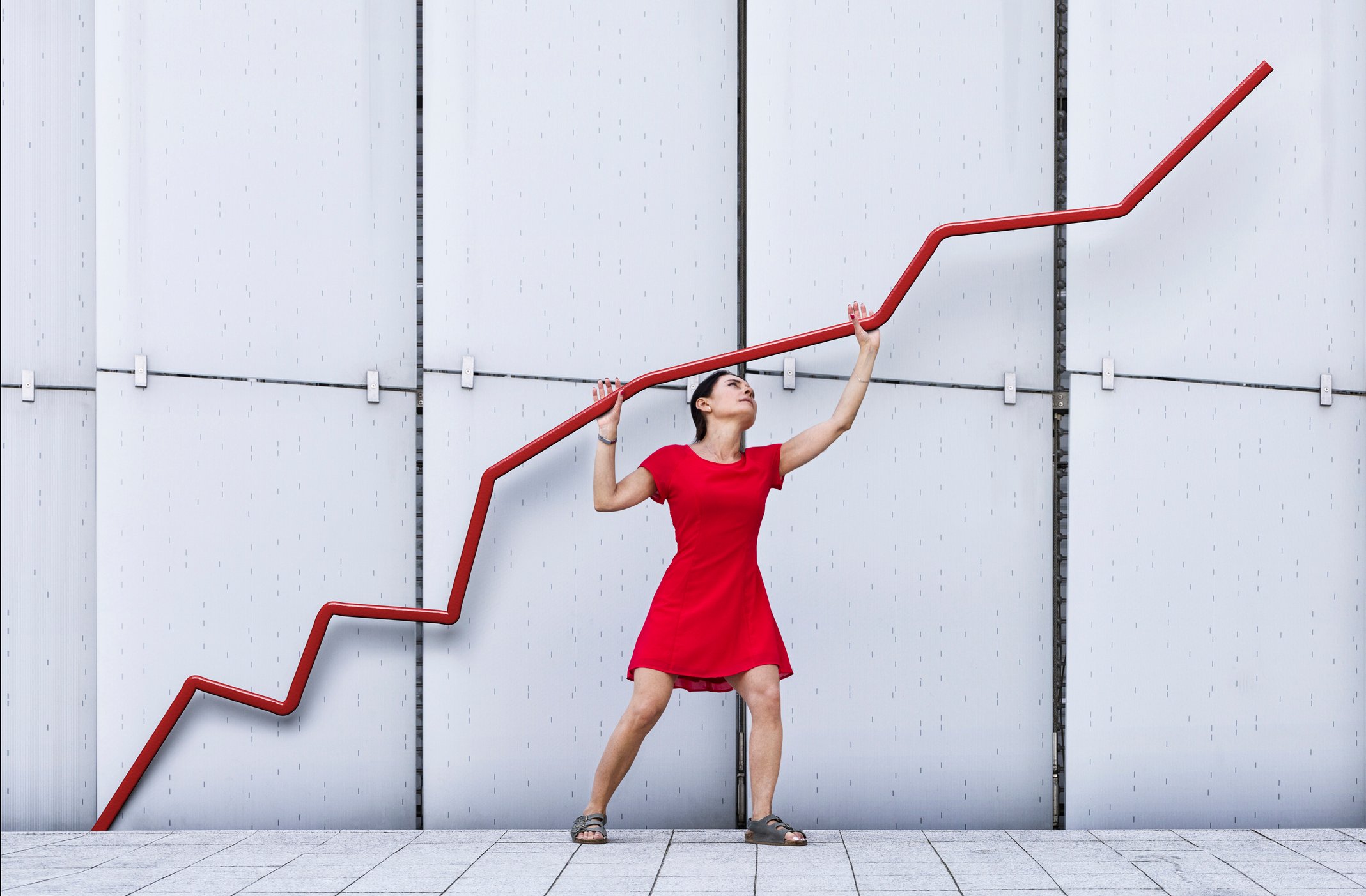 A stock chart going up with a person in a red dress standing under it and holding the line up in the sky. 