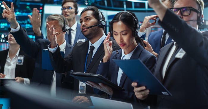 A group of financial traders and analysts actively engage on a busy trading floor.