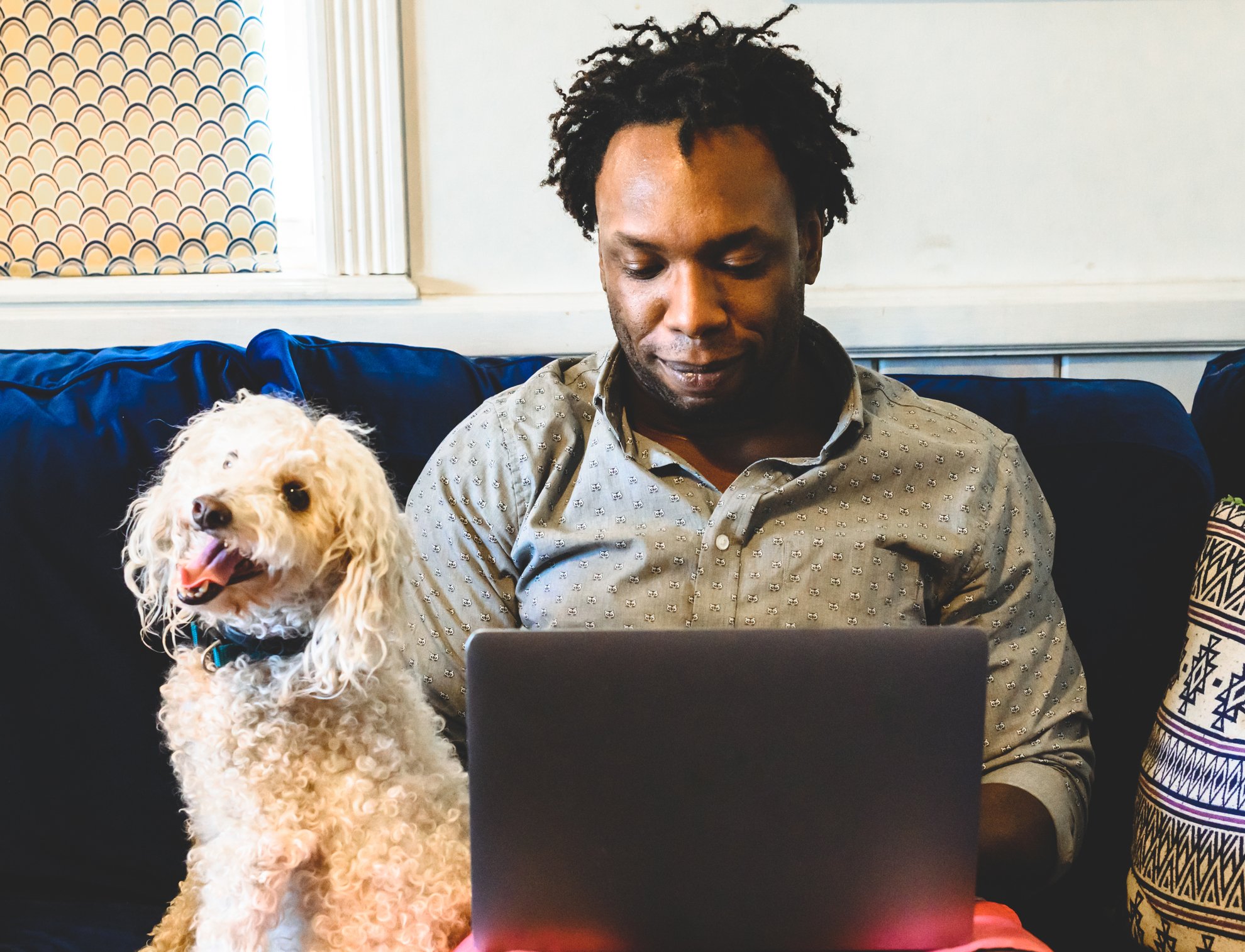 An investor sits on a couch with his computer on his lap with his little dog sitting next to him.