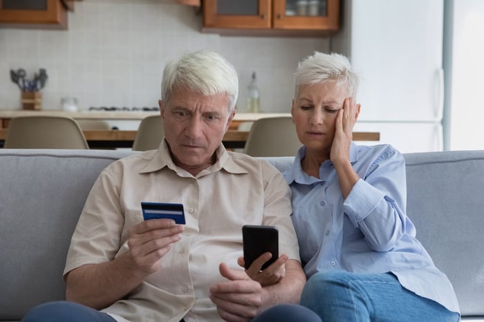 Two worried-looking people sitting on sofa and looking at phone and credit card.