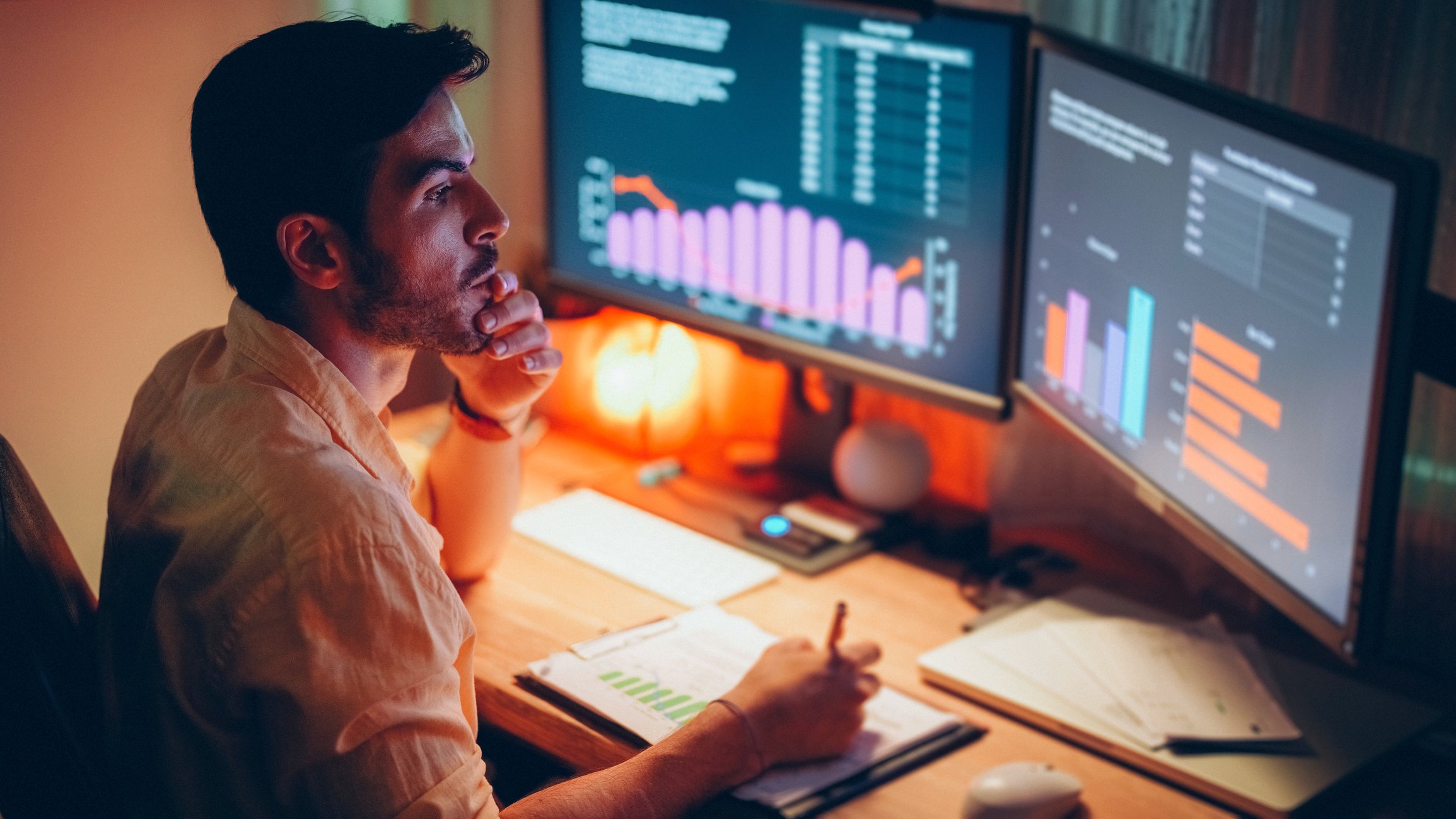 An investor writes on a printout while looking at two screens depicting stock data.