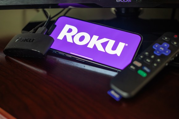 A phone showing the Roku logo sitting next to a remote control and streaming device.