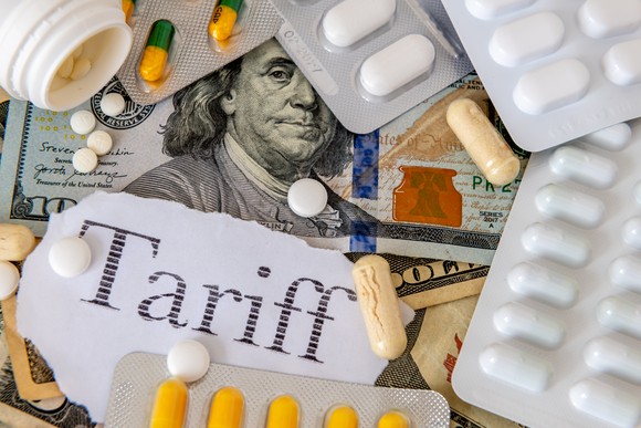 A variety of prescription pills and capsules on top of a $100 bill and a torn piece of paper reading Tariff.