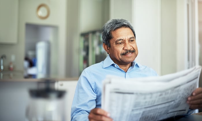 A smiling person reading a financial newspaper while seated at a kitchen table.