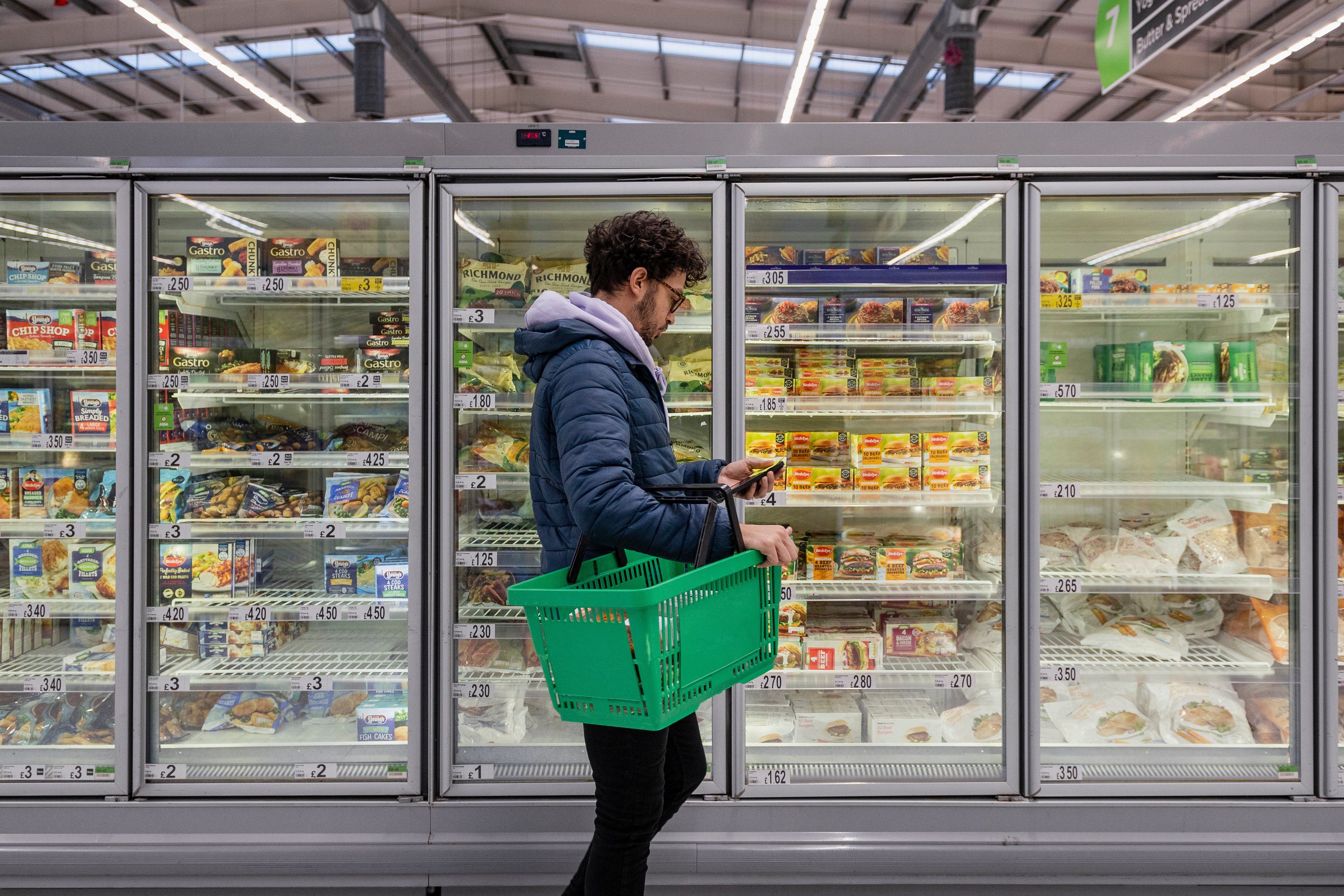 A customer holds a green shopping basket while looking at an item in the frozen foods section of a grocery store.