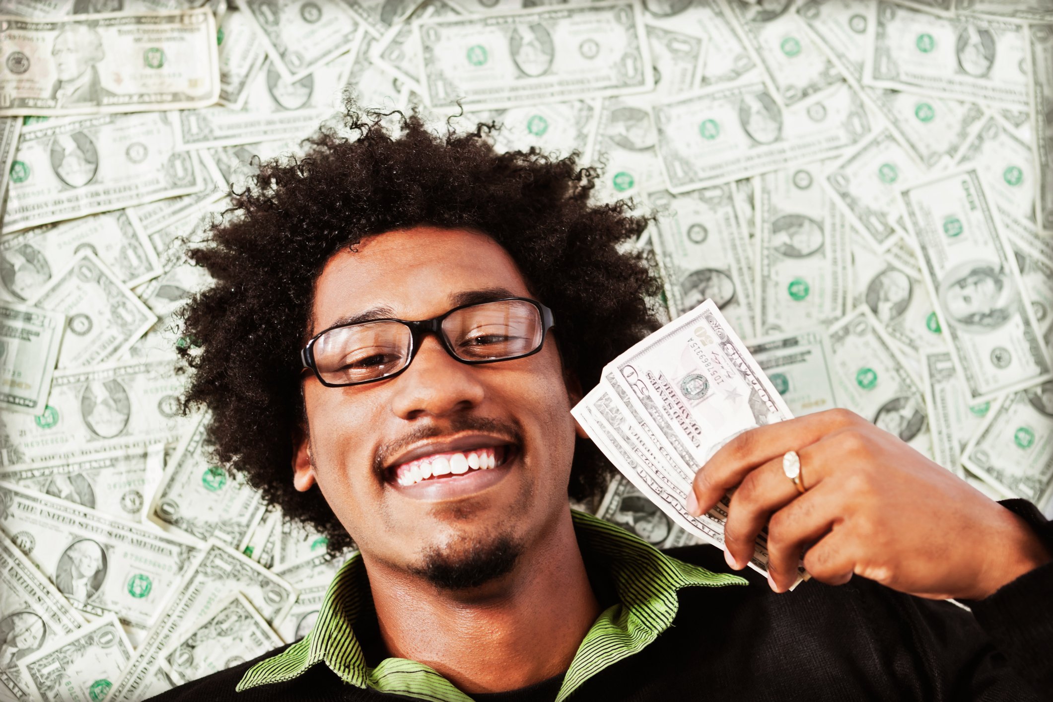A smiling person holding cash while lying on a pile of cash.