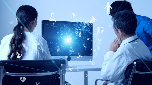 Three people looking at a monitor with icons of lab glassware, molecules, pills, and more appearing in the foreground.