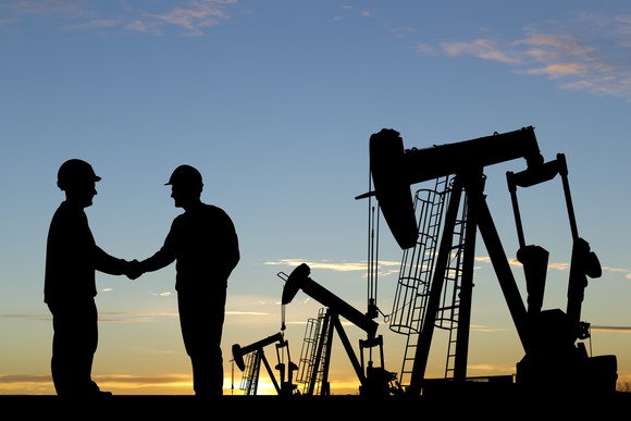 Two petroleum engineers shake hands next to a row of oil pumpjacks.