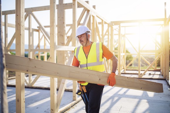 A worker carrying a board with a house under construction in the background.