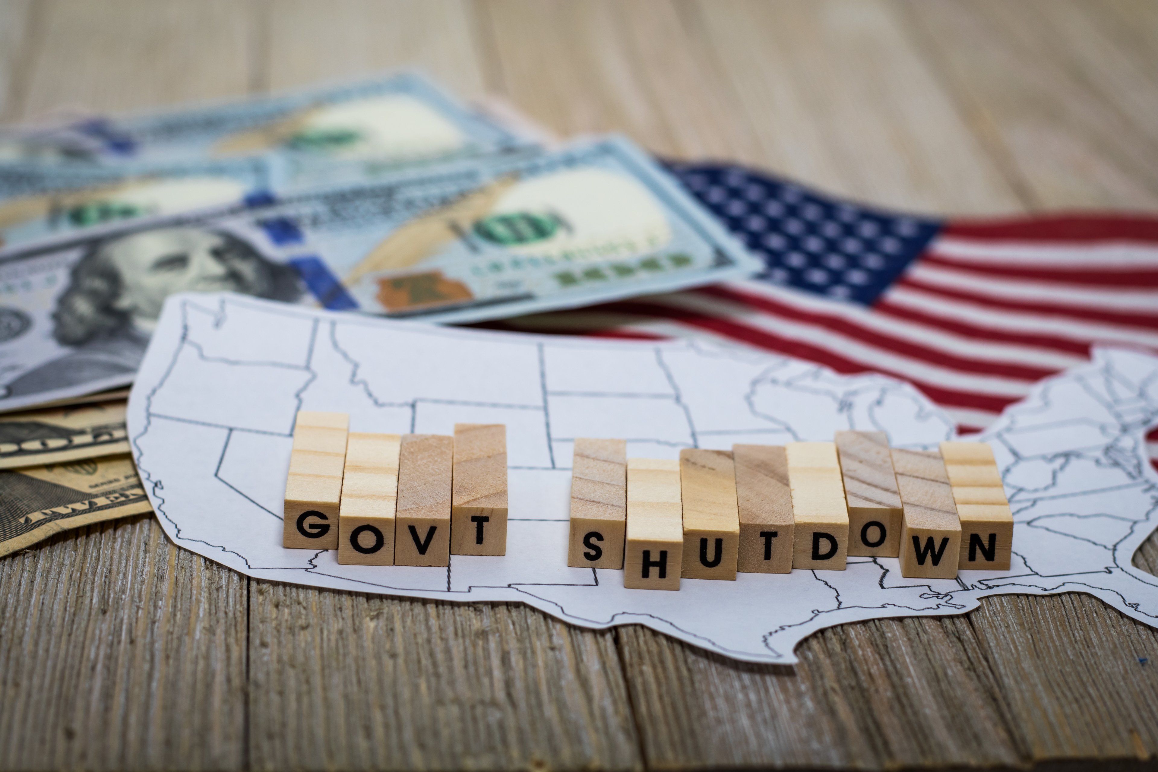 U.S. map with wooden blocks spelling “Govt Shutdown,” with dollar bills and U.S. flag in the background.