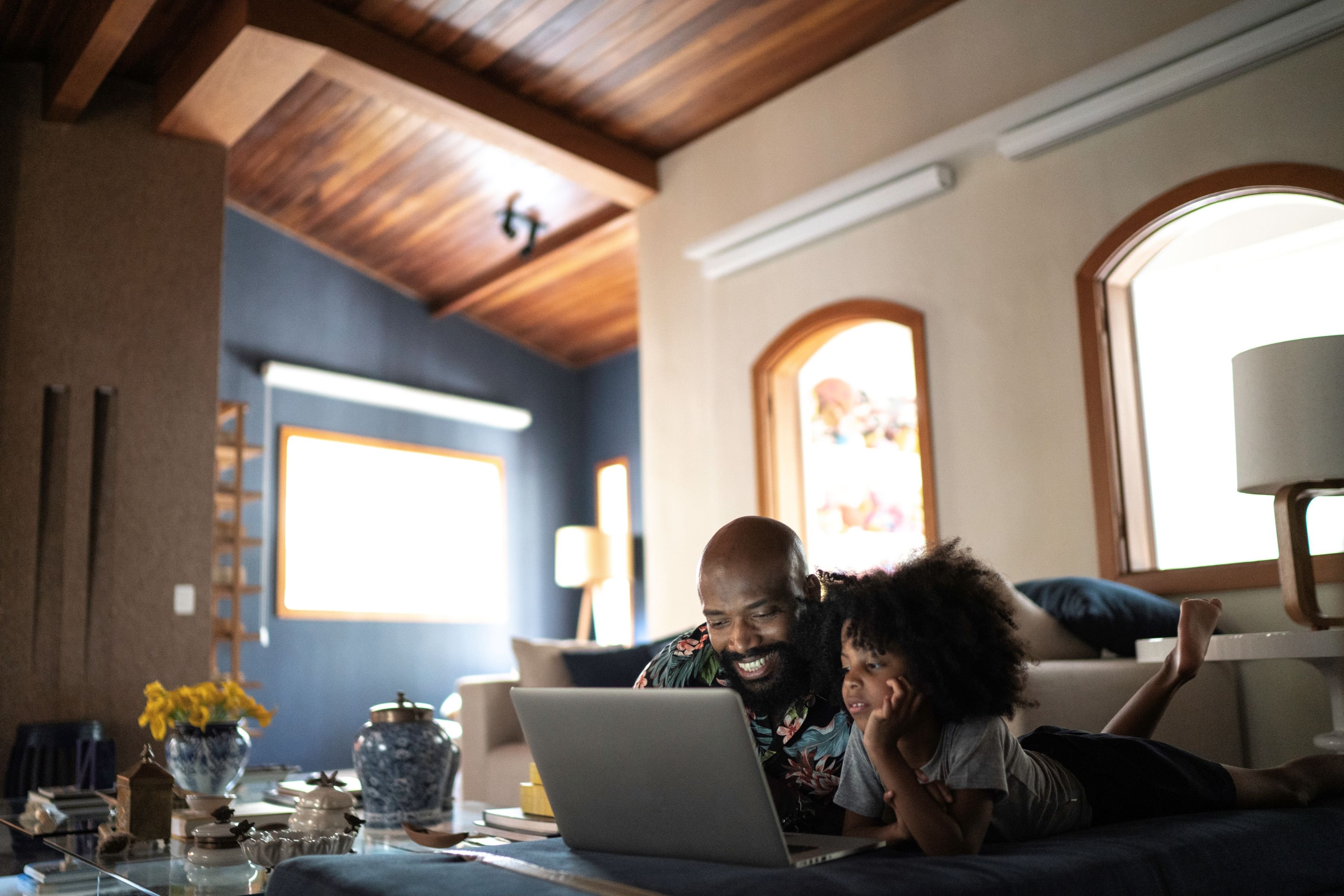 A parent helps a child while they work on a laptop in the middle of a large living room.