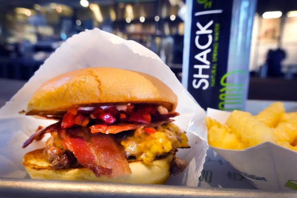A Shack Shack burger and plate of fries shown outside a store with the company logo in the background. 