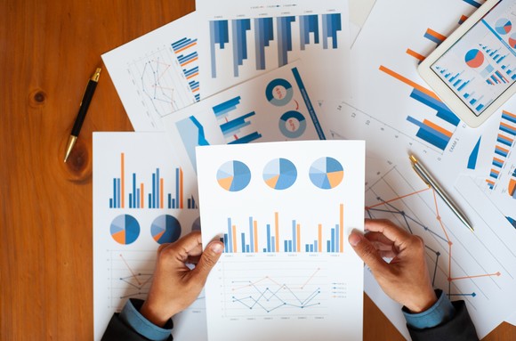 Sheets of paper with charts on them sit on a table as hands hold a sheet of paper with charts on it above them