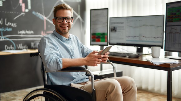 An investor sits in front of a desk with multiple computer monitors, holding a tablet.