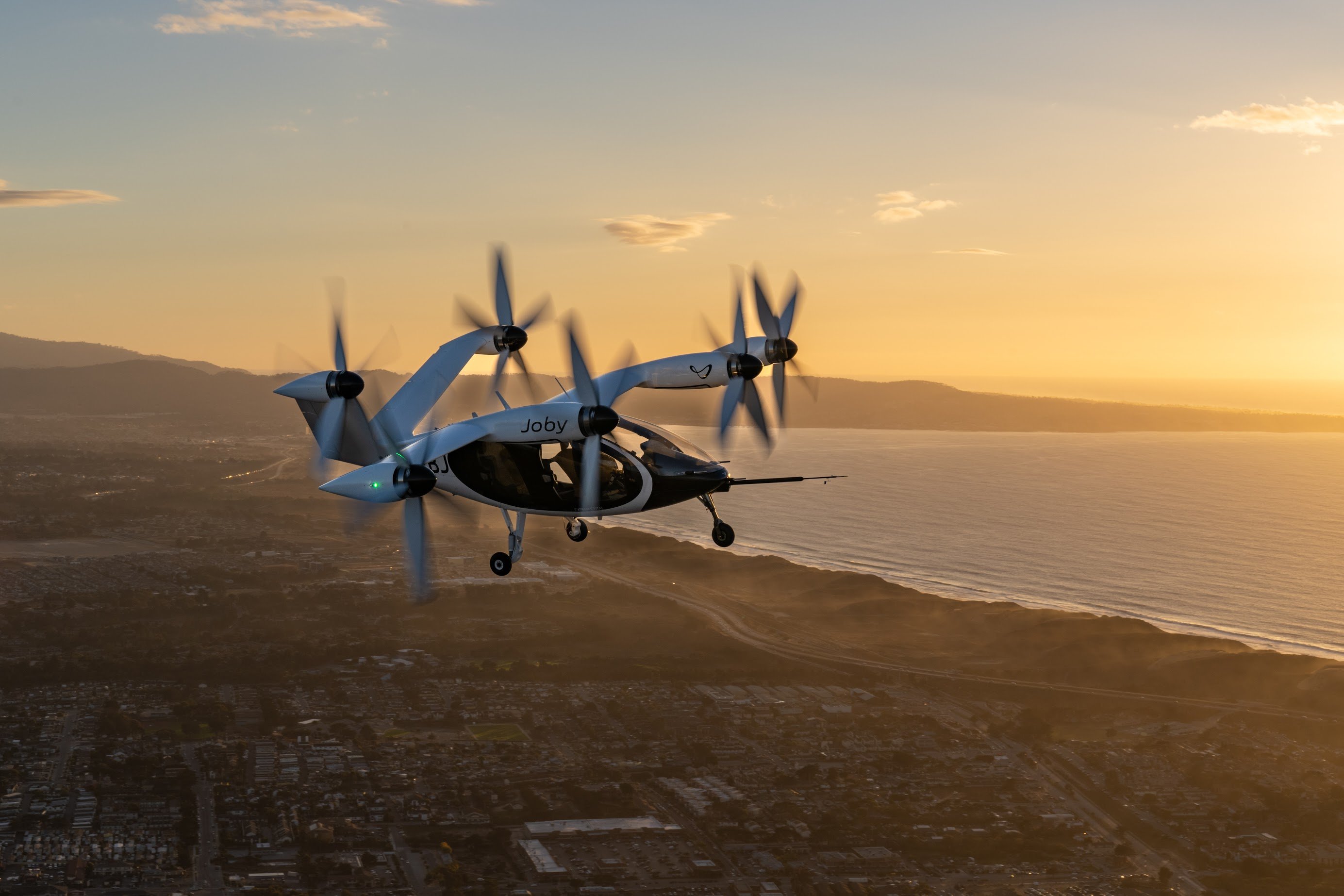 A Joby Aviation electric air taxi flying over coastal hills at sundown. 