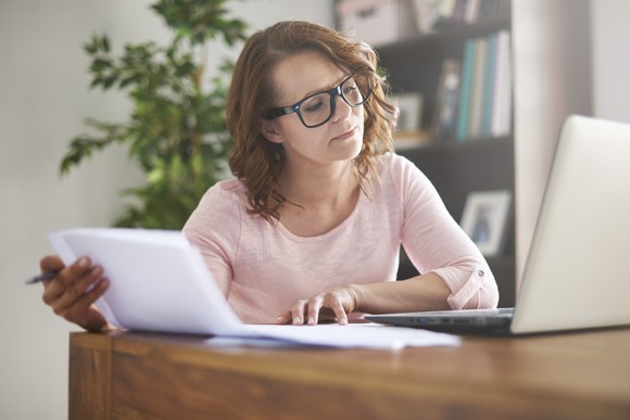 An investor holds a sheaf of papers while looking at a laptop and sitting at a desk.
