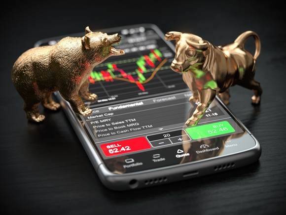 Bronze-colored bull and bear figurines face off atop a smartphone screen showing financial charts.