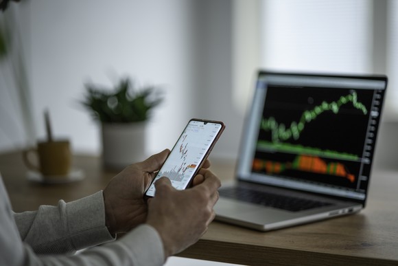 A trader trading on a phone while watching a stock price chart on a laptop screen in front of him.