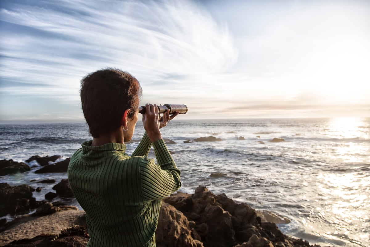 woman looks at ocean through hand held telescope.