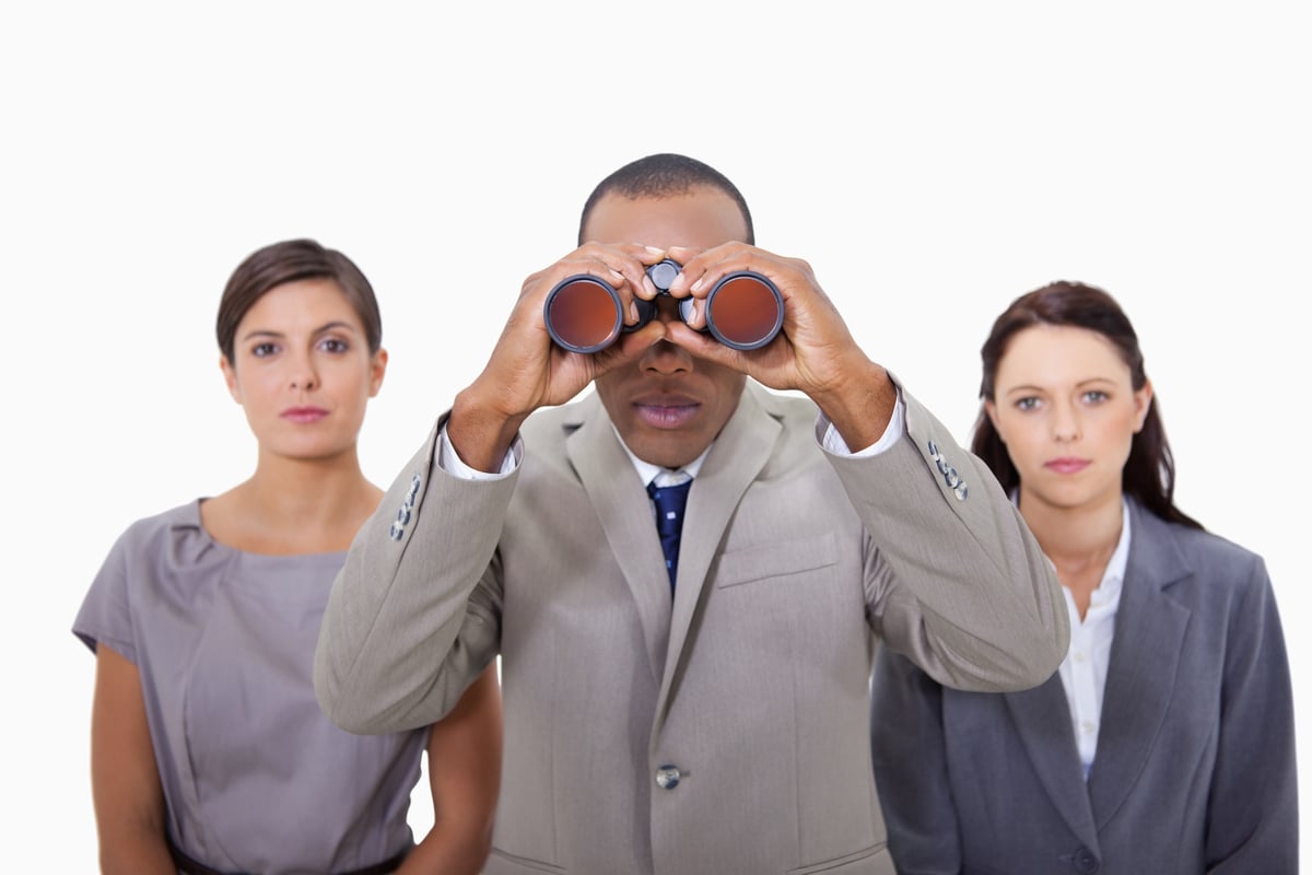 A person in a gray suit looks through binoculars, with two people standing behind.