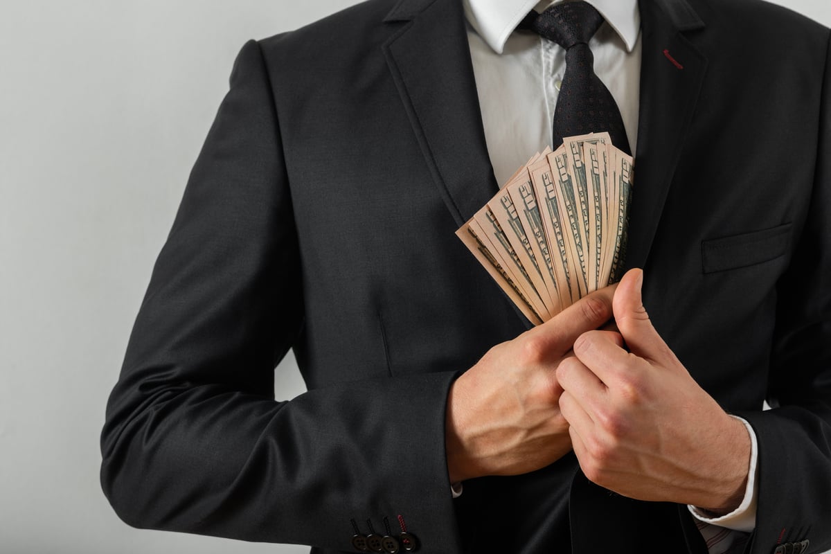 The torso of a person putting a fan of bills inside their suit jacket.