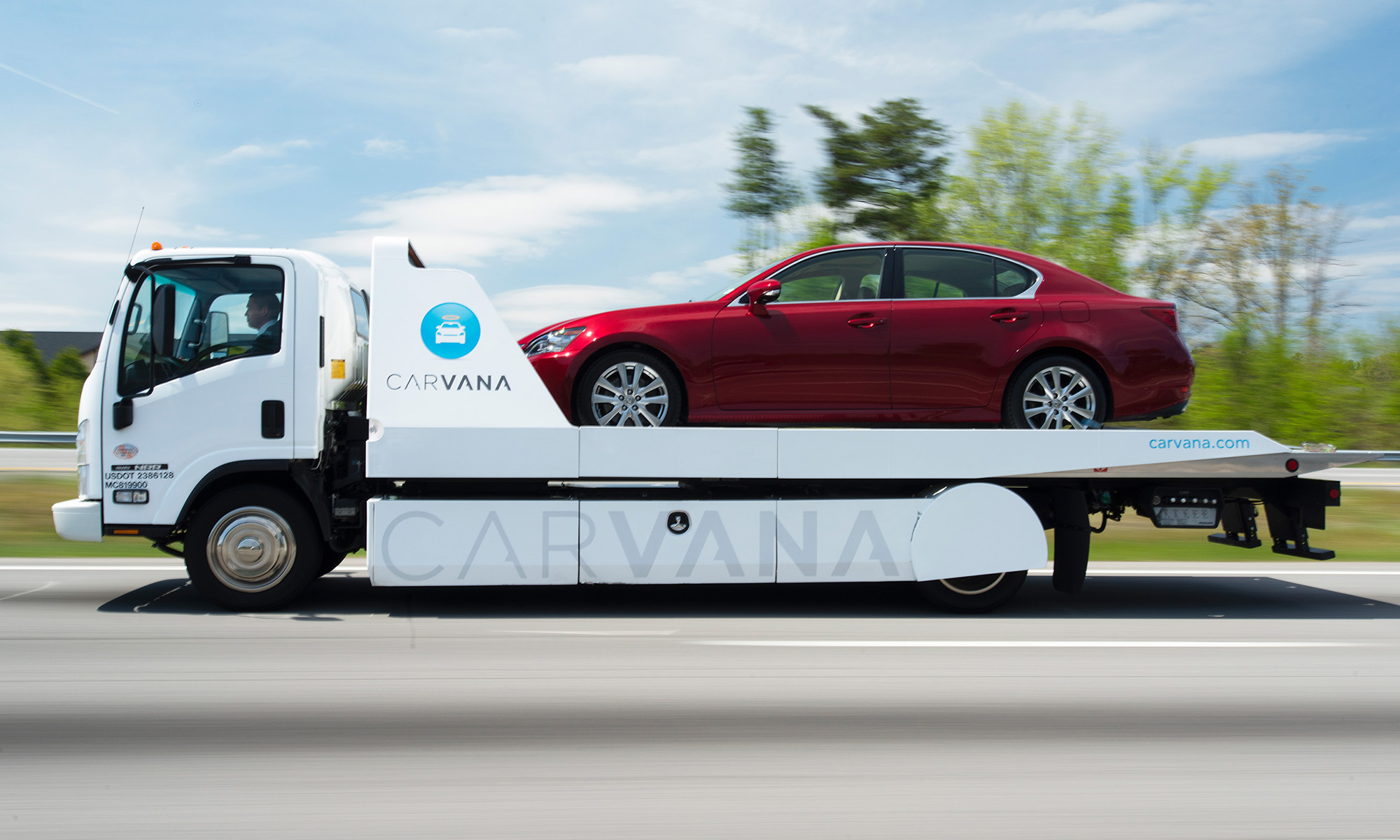 A red sedan being transported on the flatbed of a Carvana delivery truck.