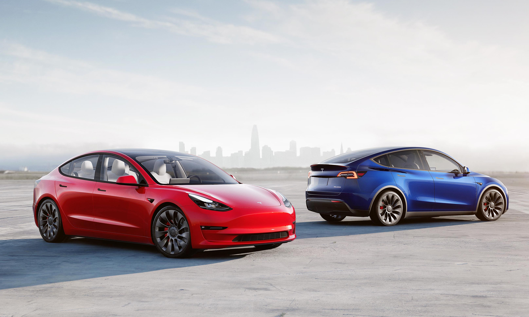 Two Tesla EVs parked on a wide, paved surface with a metropolitan skyline faintly seen in the background. 