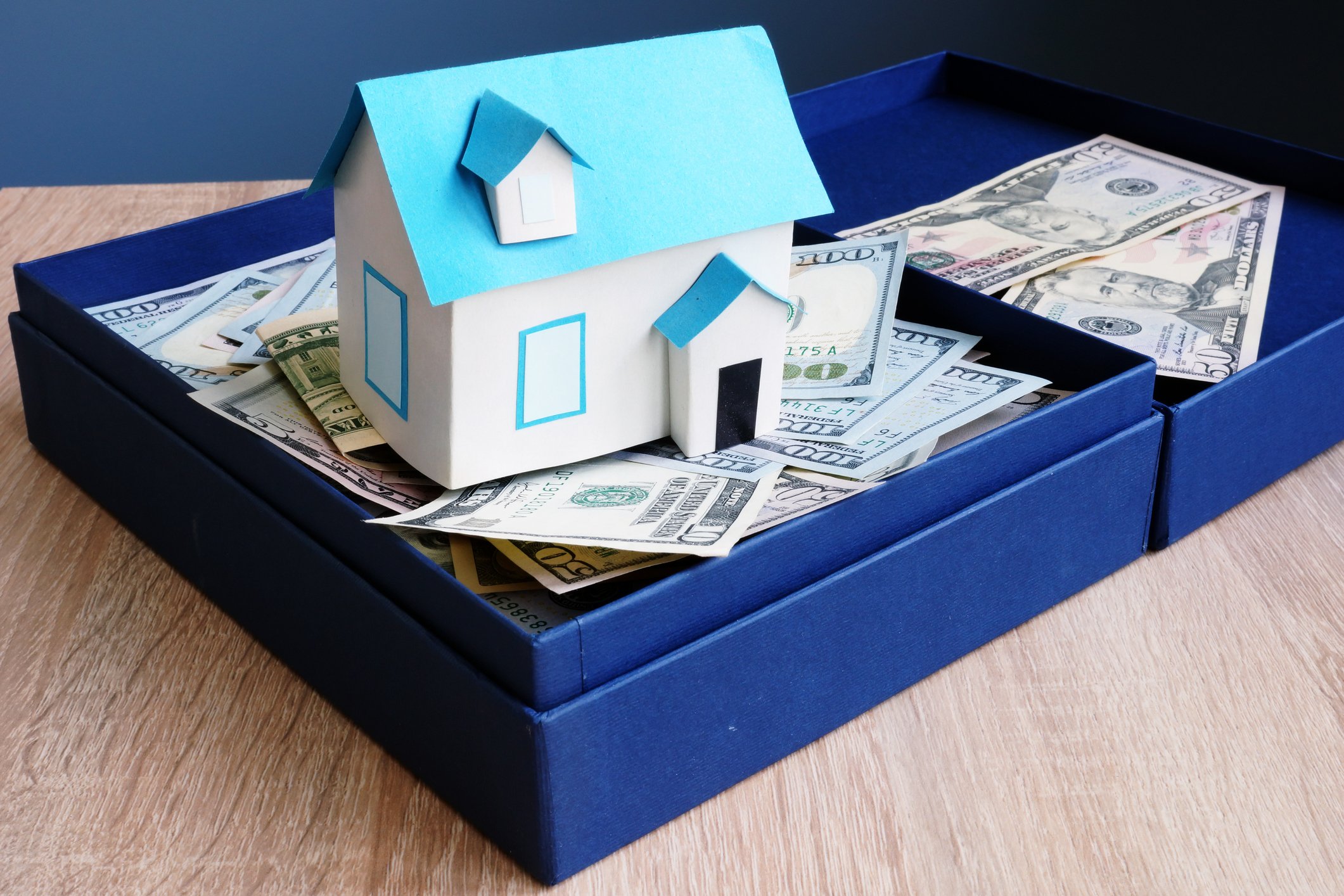 A paper model of a house sitting in a box full of $20, $50, and $100 bills