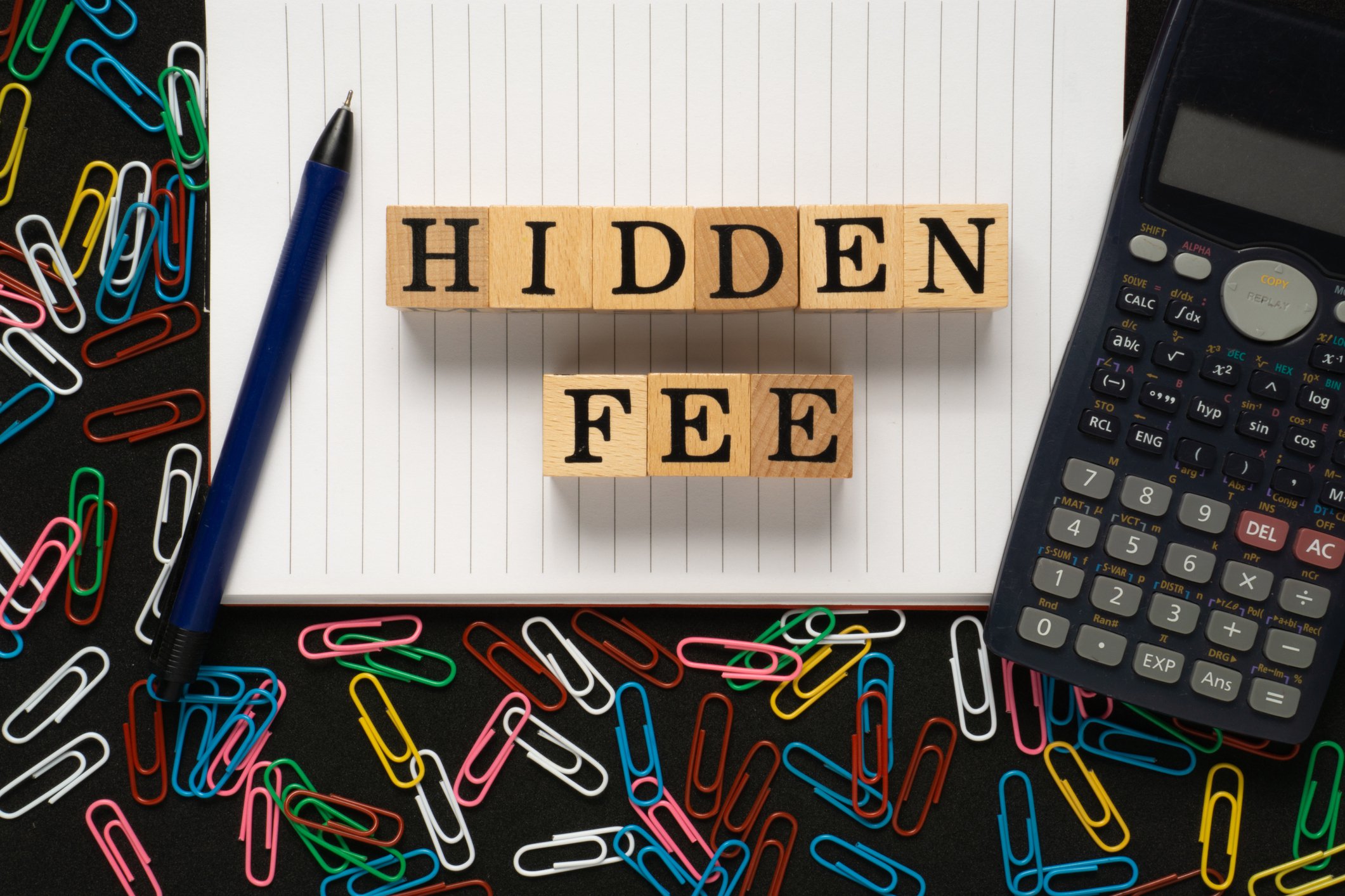 Wooden blocks spelling out Hidden Fee with pen, calculator, notebook, and paper clips nearby