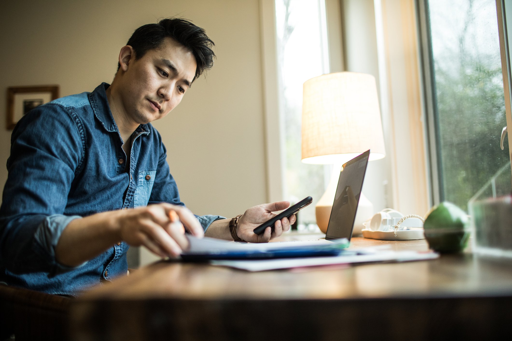 Man sits at window while working on laptop, paperwork and phone.