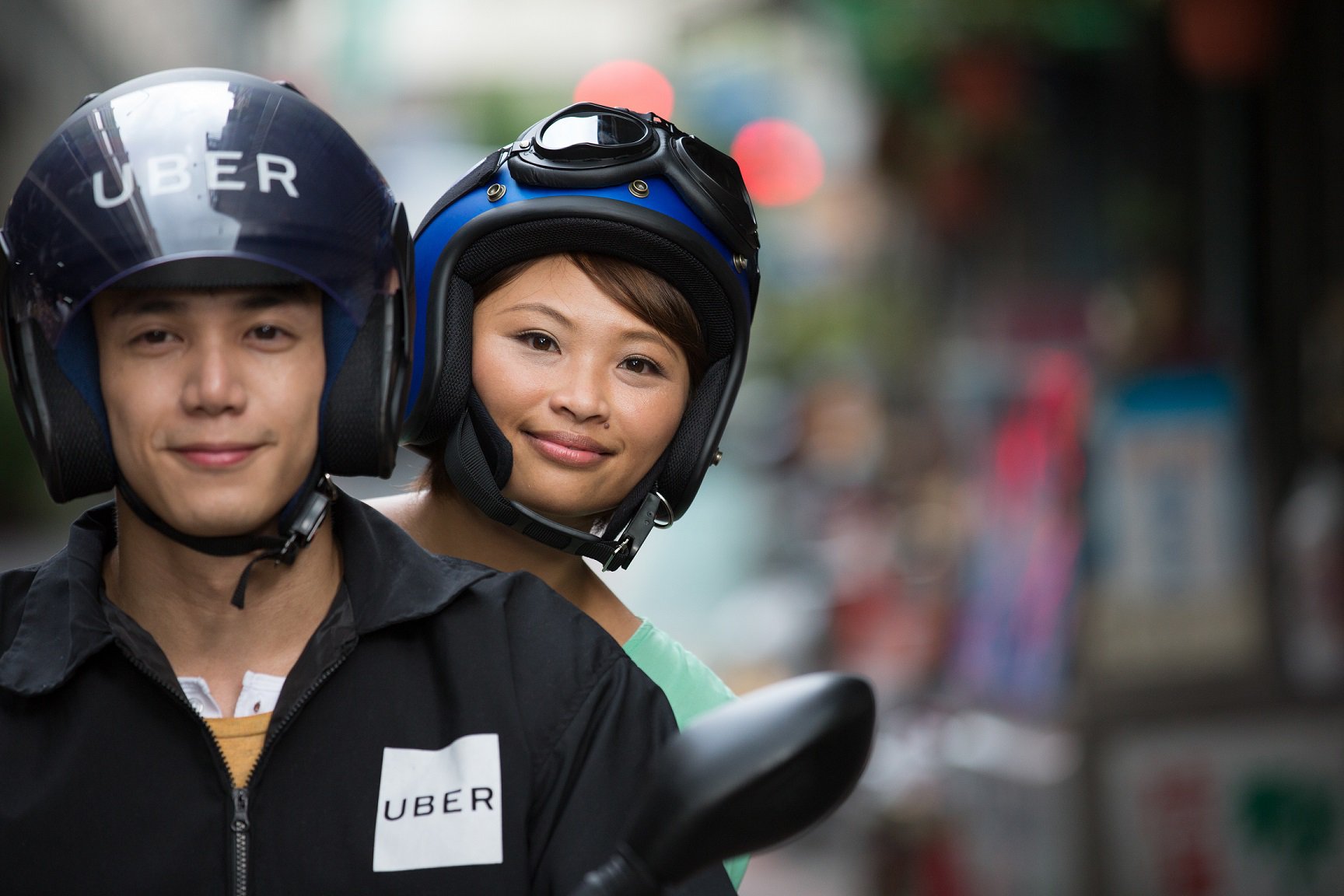 Person rides on back of motorbike behind driver with Uber helmet.