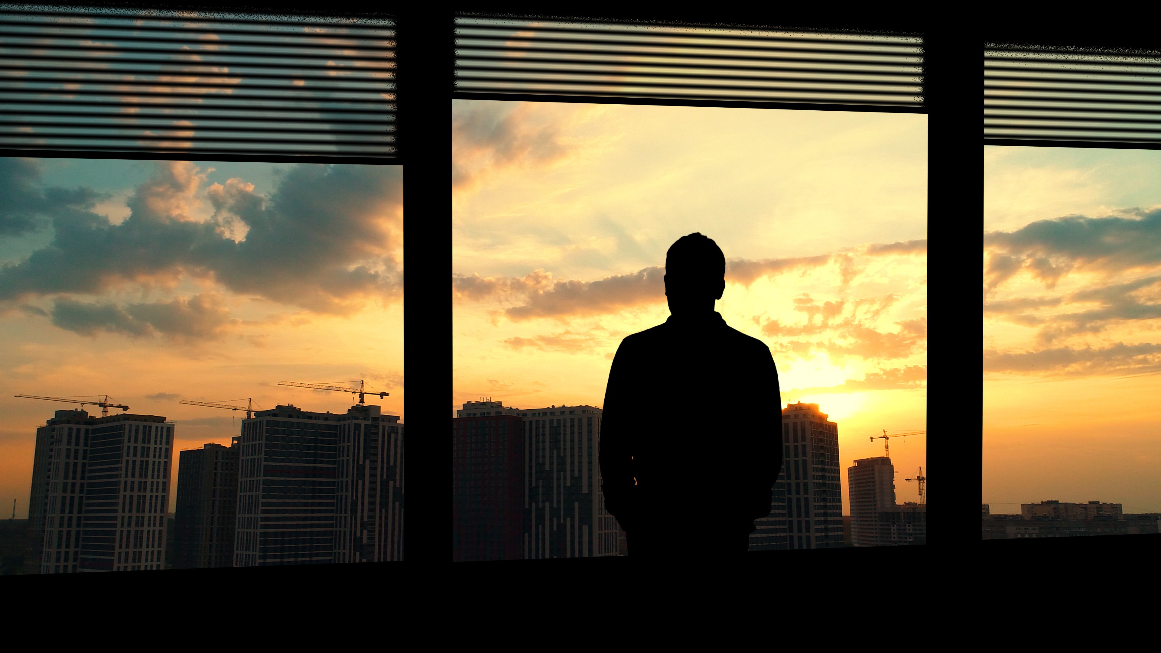 Man standing and looking out window at sunset over city.