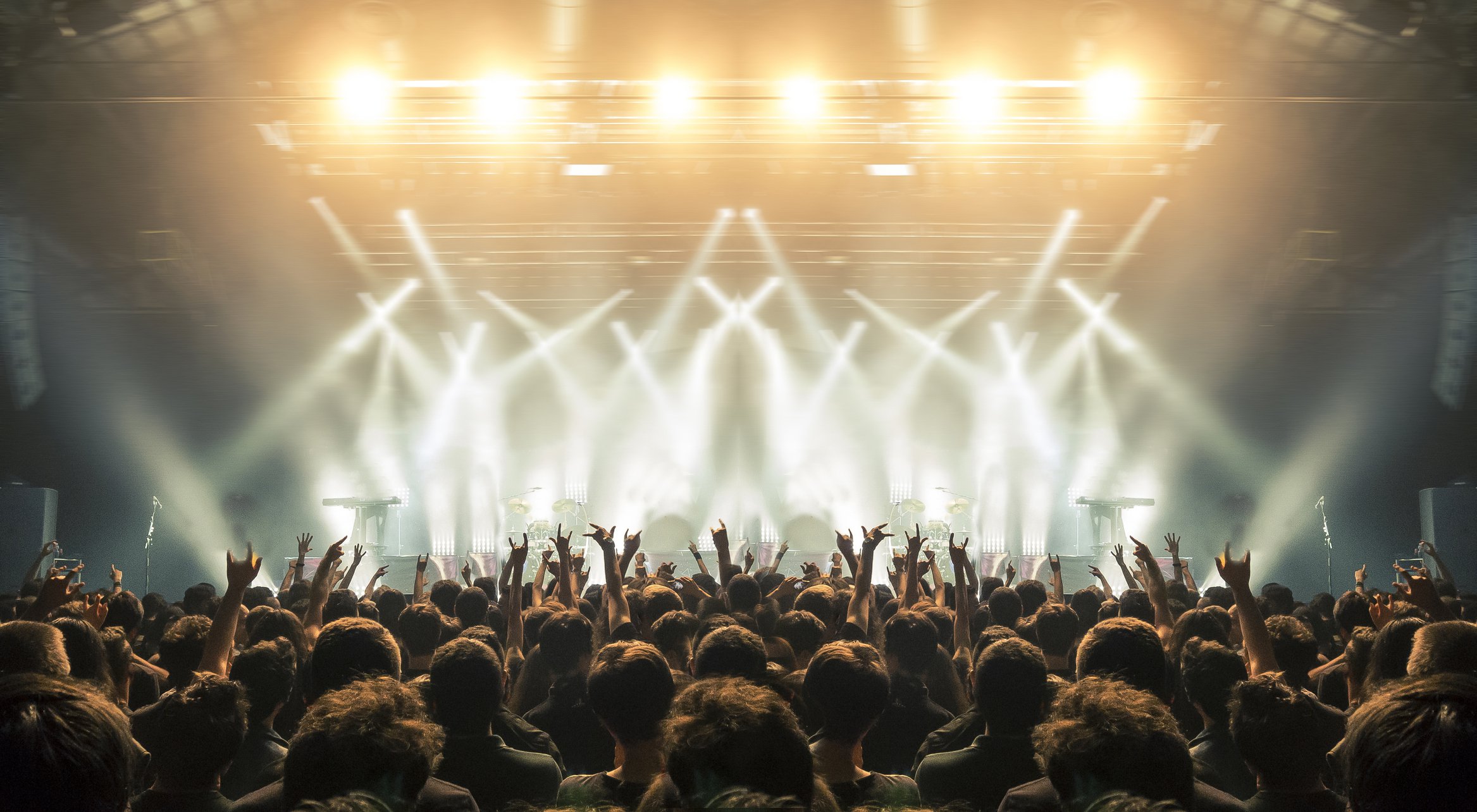 A crowd with hands in the air backlit by stage lights at a concert.