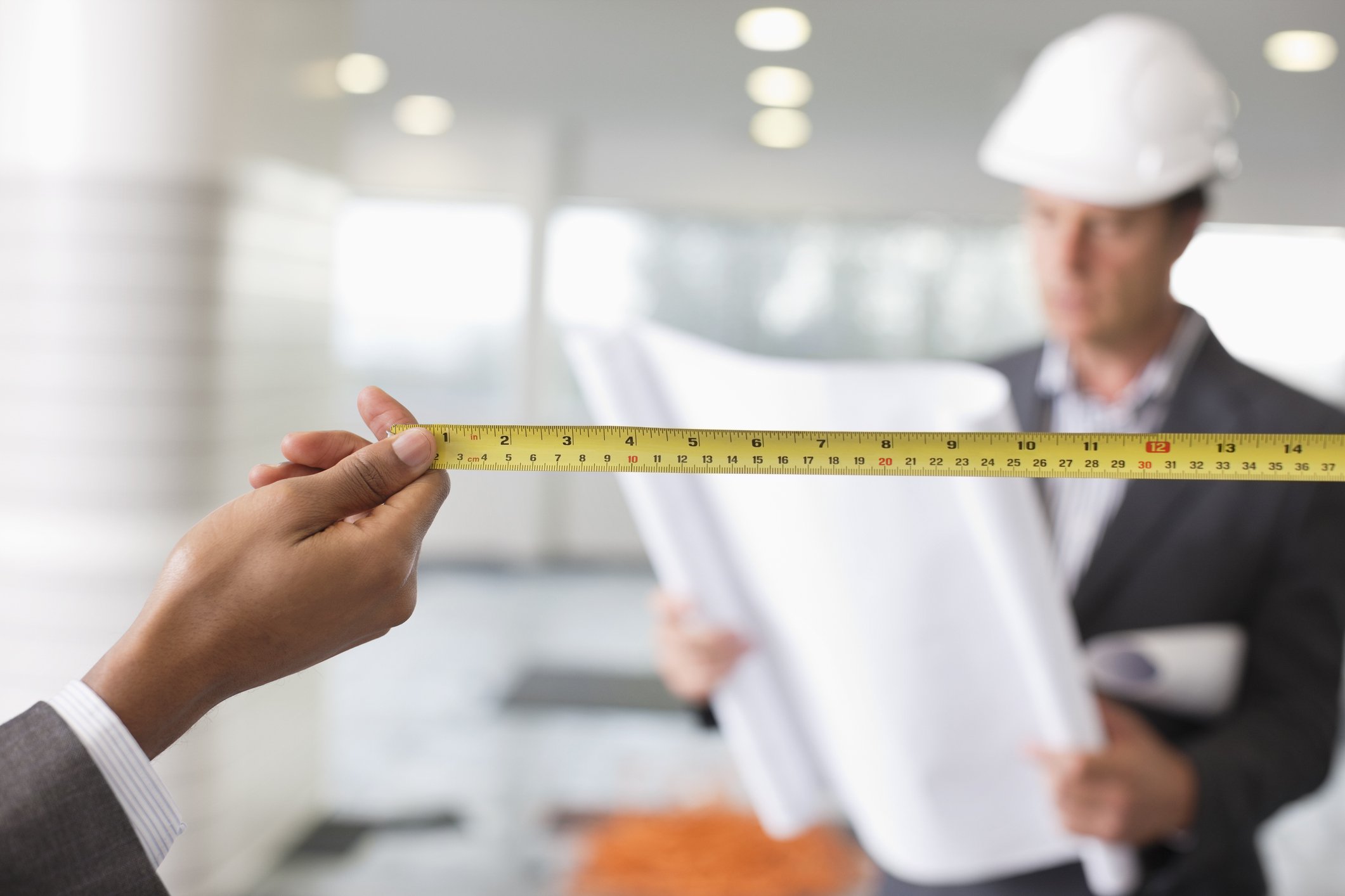 Person holding a tape measure in a blank space and construction worker in background.