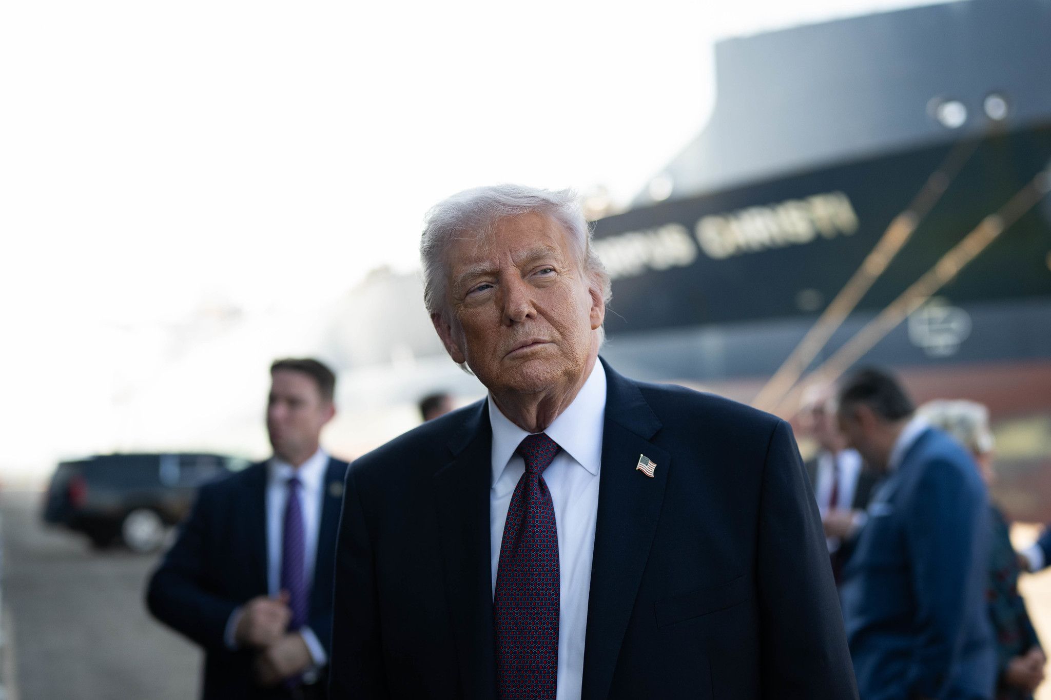 Donald Trump is preparing to deliver remarks, with a large shipping vessel in the background.