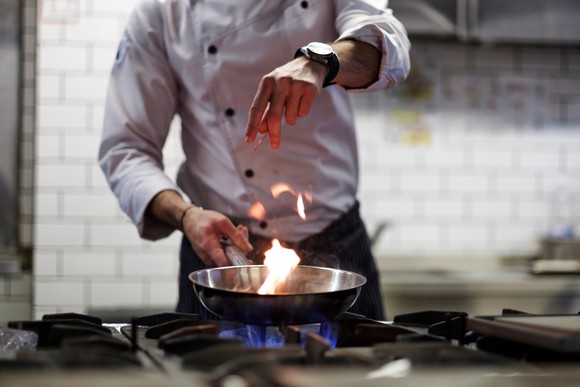 A chef drops ingredients into a saucepan on a stovetop.