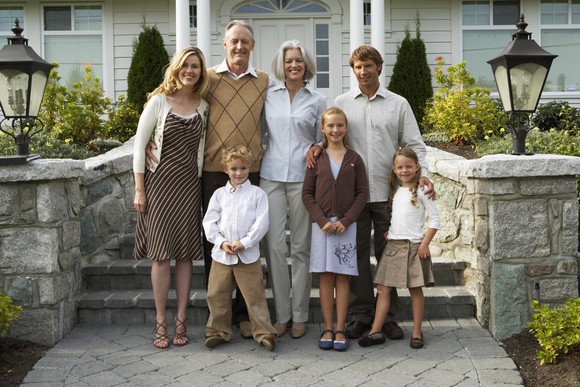 Three generations of a white, middle-class family stand in front of their home