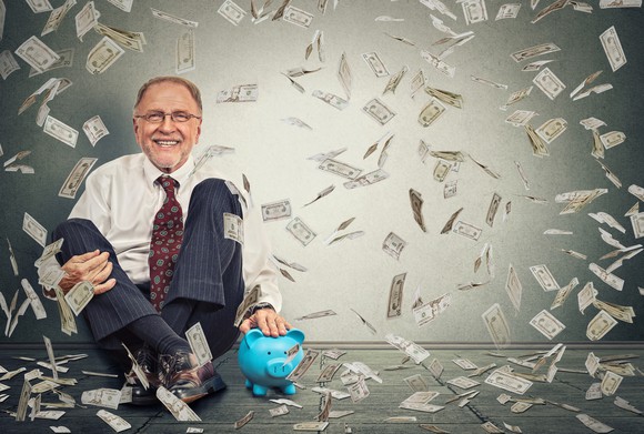 An older businessman sits with his back against a wall as money falls down around him and a piggy bank sits on the floor beside him.