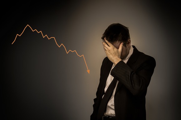 A businessman stands in front of a declining arrow, holding his head in his hands.