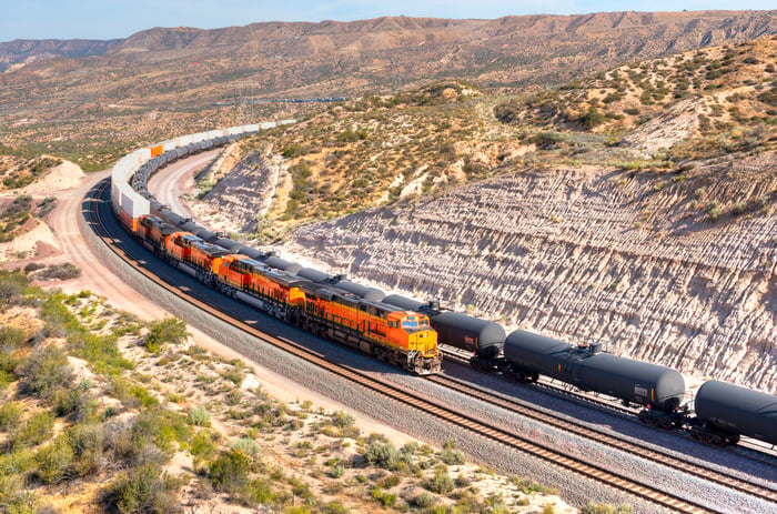 A long train loaded with double-stack white cargo containers winds its way around tight s-curves in mountain countryside.