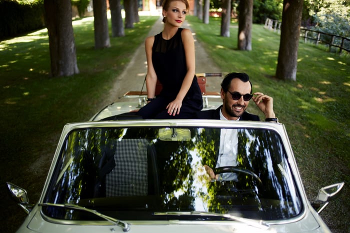 Rich young couple in convertible, with guy in sunglasses and woman perched on back seat